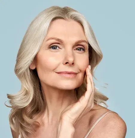 A woman with shoulder-length blonde hair gently touches her face. She is wearing a light-colored camisole, against a light blue background.