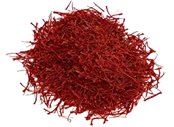 A pile of dried crimson saffron threads rests on a white background; a small, dark red cylindrical object is visible to the right.